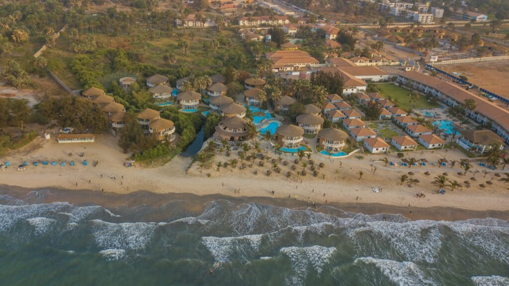Panoramic drone view of Balafon Beach Resort on Kololi Beach, featuring round villas, palm-lined pools, and pristine beachfront. A premier destination for relaxation and beach getaways in ...